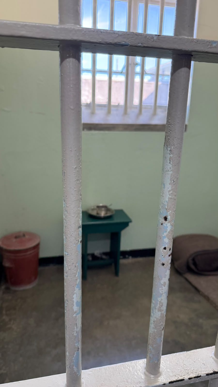 Interior da cela de Nelson Mandela em Robben Island, vista através das grades: chão de cimento, um cobertor dobrado, uma mesinha verde com uma caneca de metal e uma janela gradeada.