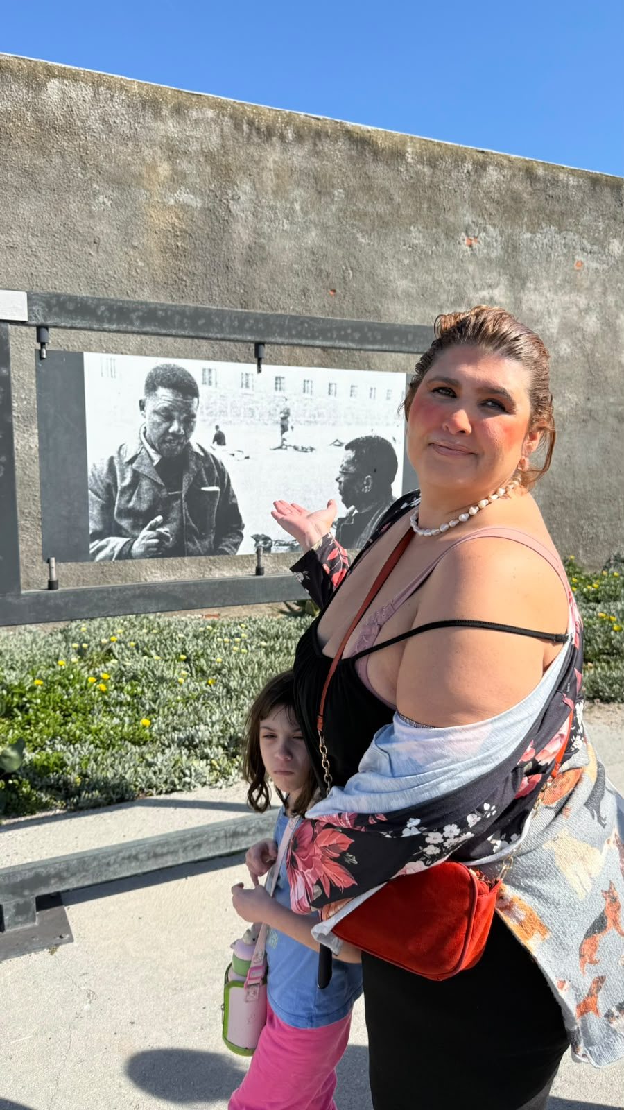 Diana e a filha diante de um painel com uma fotografia em preto e branco de Nelson Mandela no pátio da prisão.