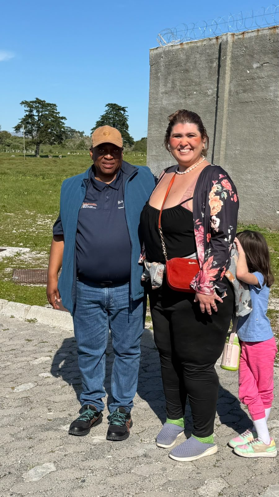Sello Motse, guia de Robben Island e ex-prisioneiro político, ao lado de Diana e da filha, em frente a um muro do presídio.