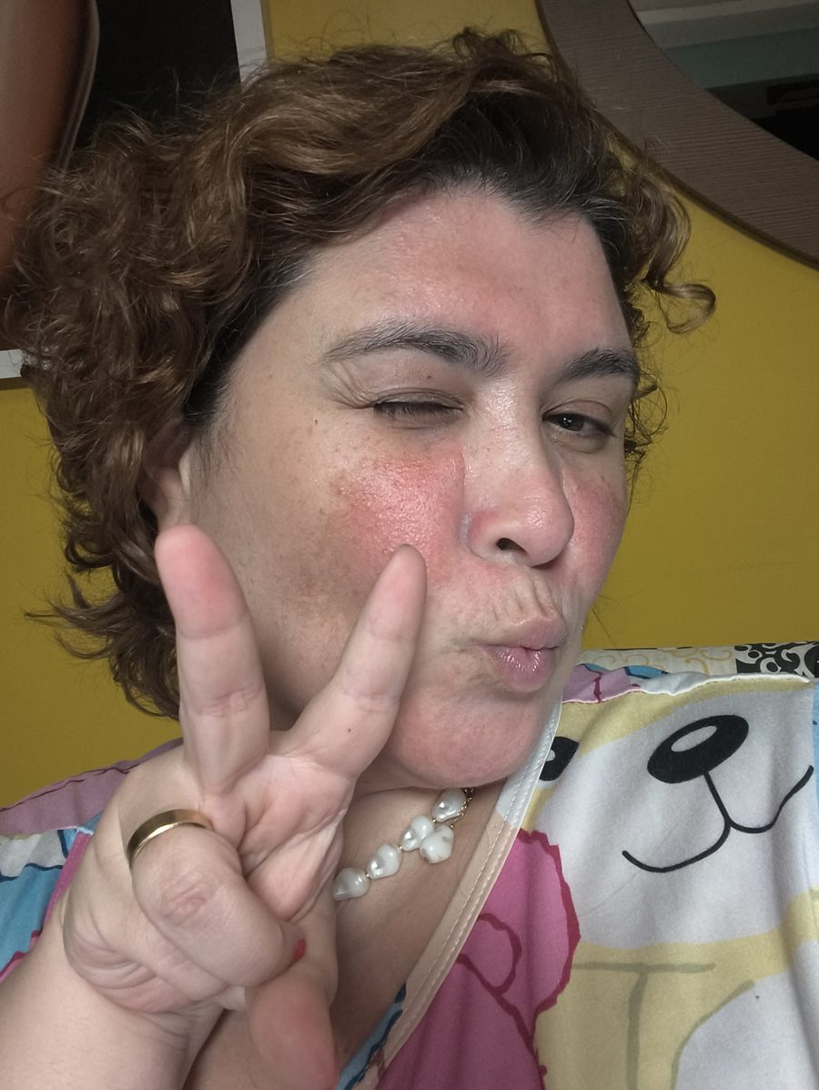 Selfie da Diana piscando um olho e fazendo V com os dedos, tirada de madrugada com luz quente e fraca na sala.