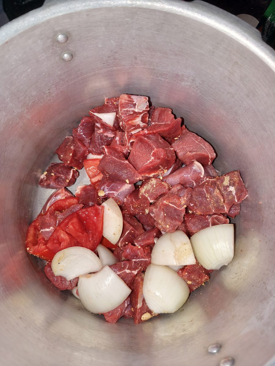 Panela de pressão vista de cima, cheia de cubos de músculo bovino crus, pedaços de tomate e cebolas inteiras descascadas, sem água nem óleo.