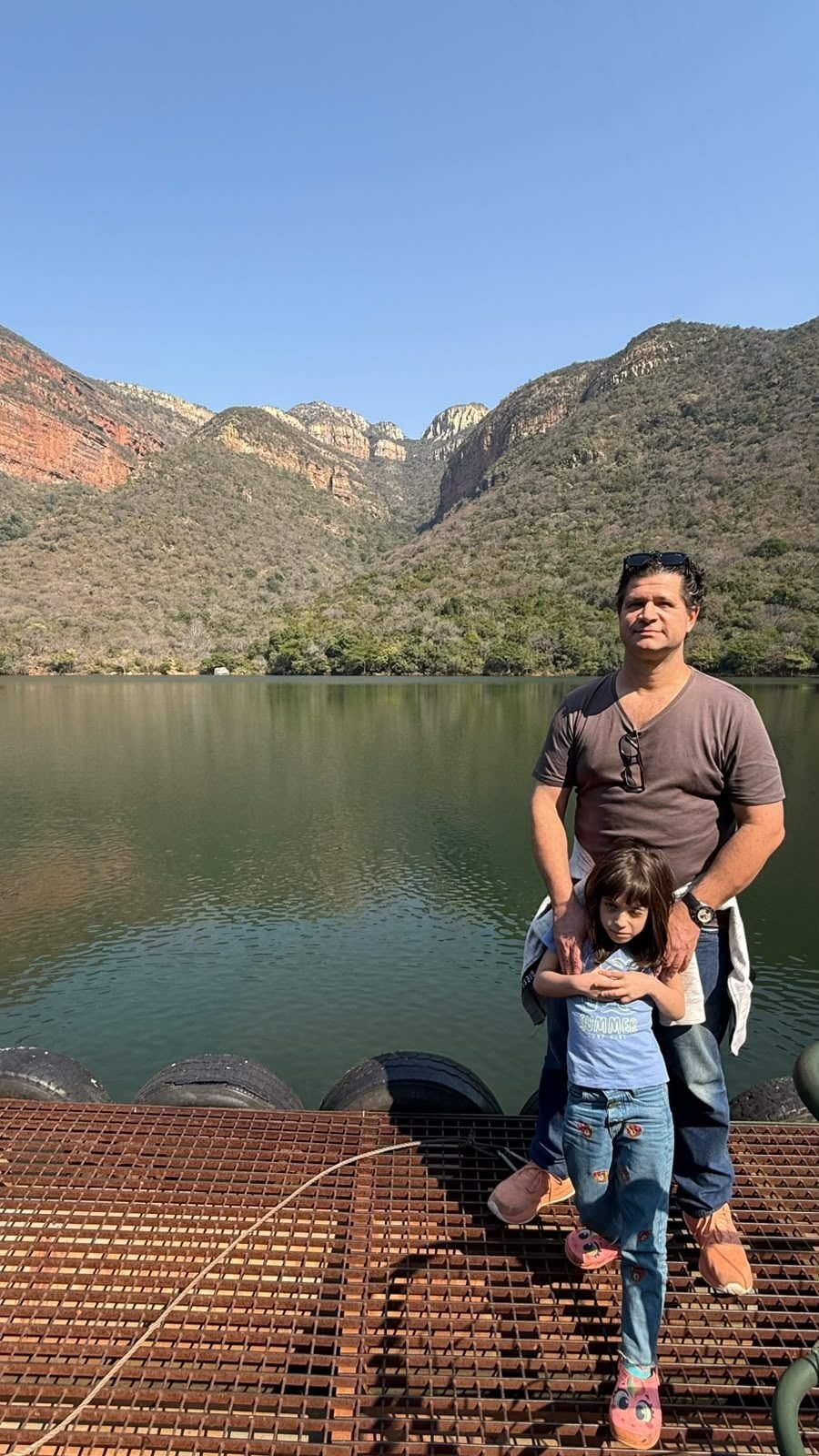 Nicco e Dinah em pé no atracadouro flutuante da represa do Blyde River Canyon, com o cânion ao fundo e nenhum barco na água