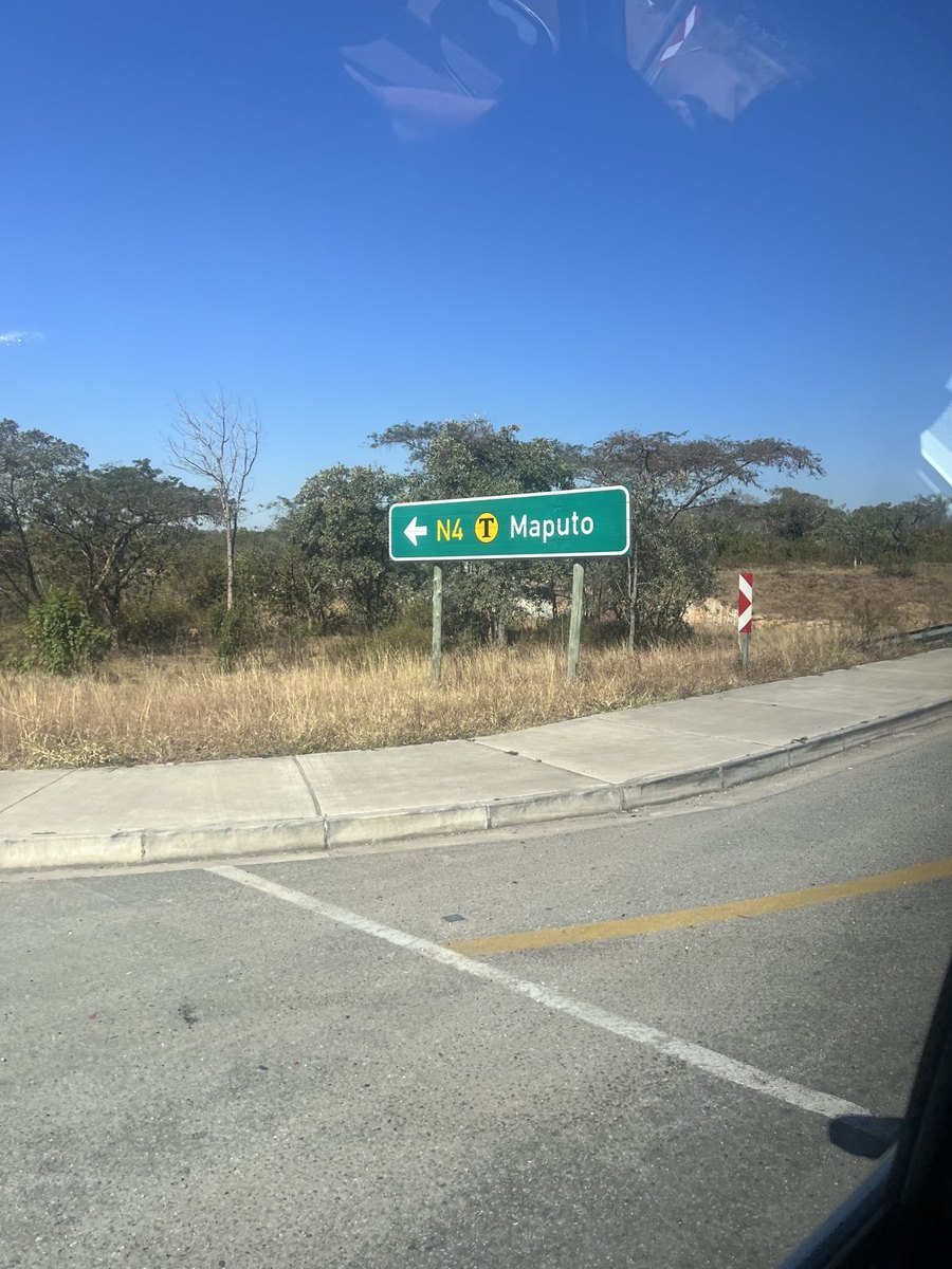 Placa verde de estrada na beira da pista indicando o caminho para Maputo, com mato seco e céu azul em volta.