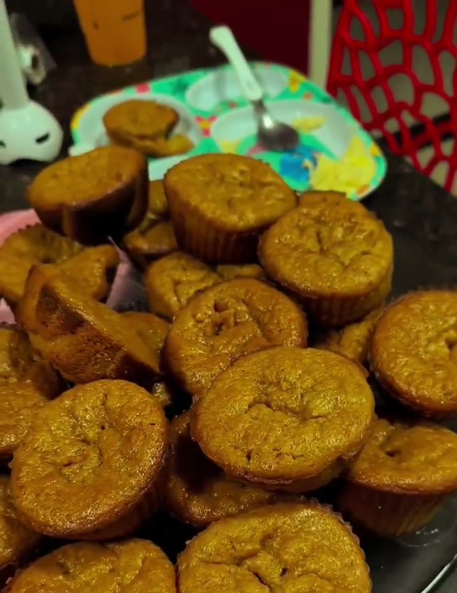 Boleira preta cheia de bolinhos de banana assados e dourados, com o prato infantil colorido da filha ao fundo, já com um bolinho servido.