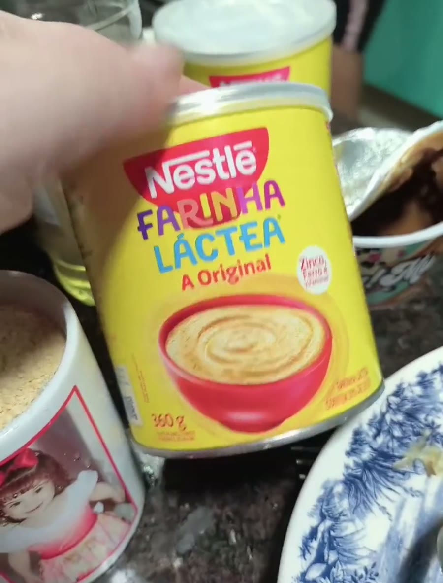 Lata amarela de farinha láctea Nestlé A Original, de 360 g, segurada sobre a bancada da cozinha, ao lado de uma caneca com a farinha já medida.
