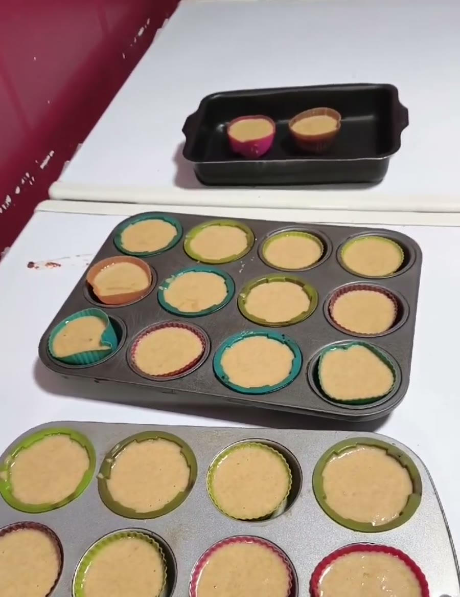 Duas formas de cupcake com forminhas de silicone coloridas em formato de flor, cheias da massa crua do bolo de banana, apoiadas sobre o fogão branco.
