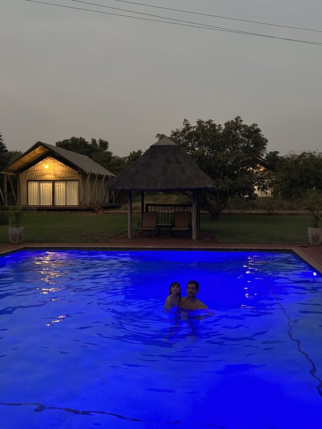 Chalé de lona e sapé iluminado ao entardecer, com um quiosque de sapé ao lado e a piscina acesa em azul, onde estão o Nicco e a Dinah