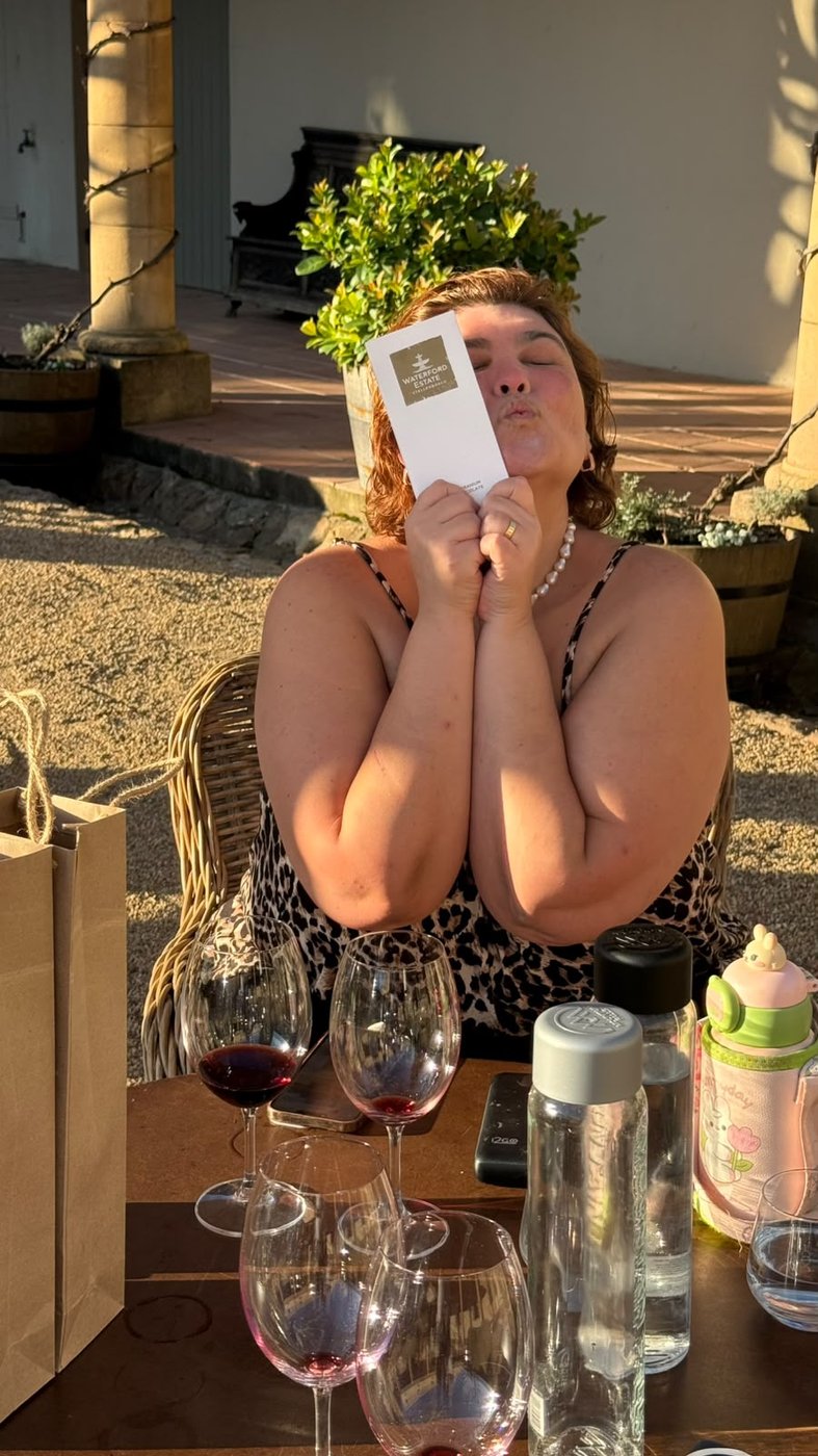 Diana sentada à mesa do Waterford na luz do fim de tarde, de olhos fechados, beijando a embalagem da barra de chocolate segurada com as duas mãos