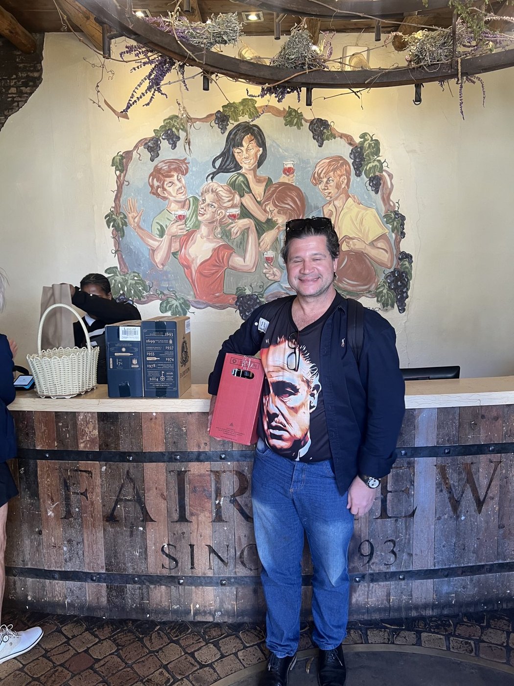 O Nicco em pé no balcão da Fairview, feito de aduelas de barril, com a caixa de vinho na mão e o mural pintado atrás