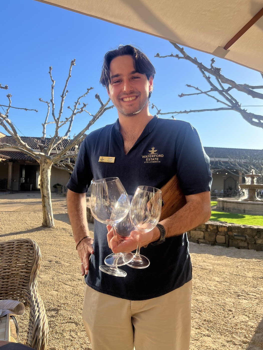 Liam, de camisa do Waterford Estate, no pátio da vinícola segurando três taças e a tábua de pareamento
