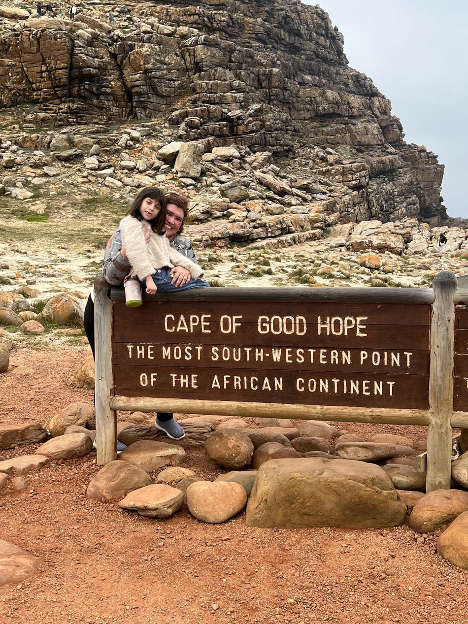 Foto da família junto à placa de madeira do Cabo da Boa Esperança, com o paredão de rocha ao fundo