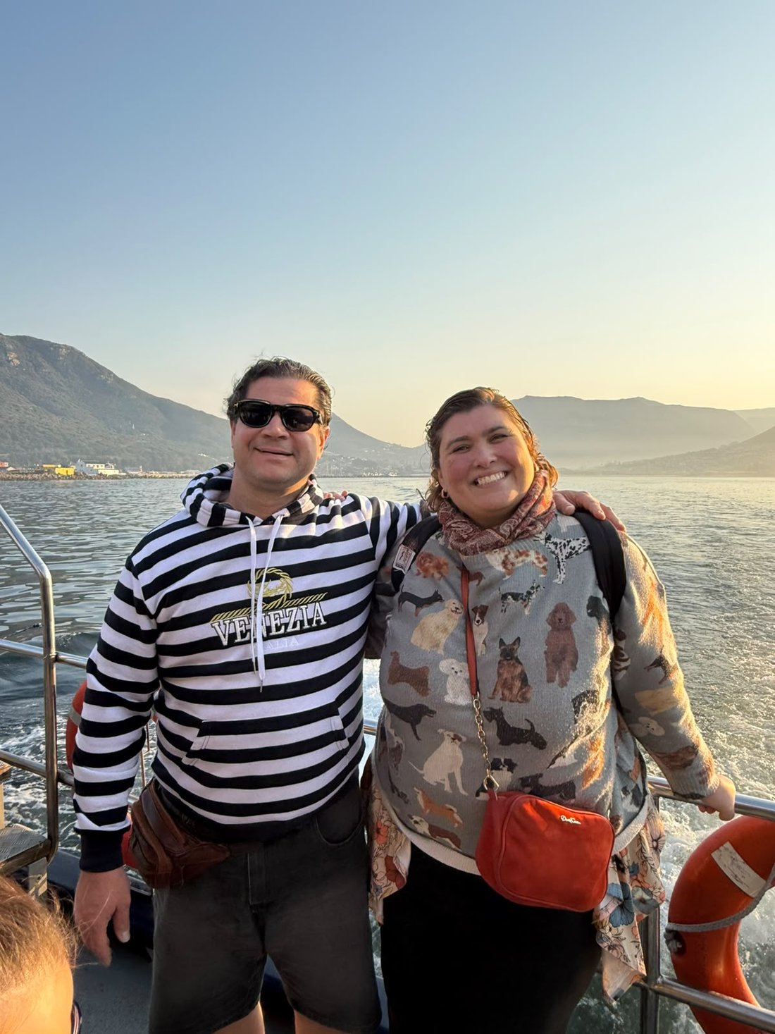 Diana e Nicco abraçados no convés do barco, com a baía de Hout Bay e as montanhas ao fundo