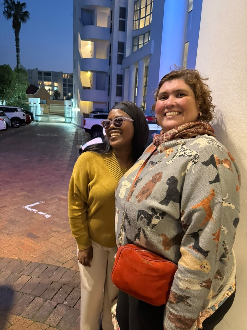 Nana e Diana lado a lado no fim da tarde em Sea Point, sorrindo, com os prédios acesos ao fundo