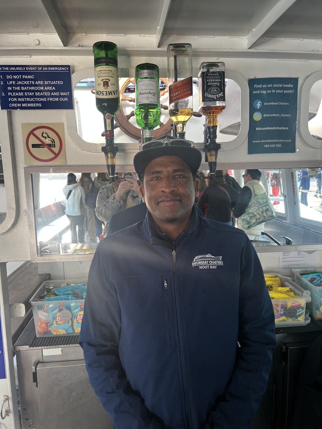 Ricky, tripulante da Drumbeat Charters, de boné e casaco da empresa, em pé na copa do barco em Hout Bay