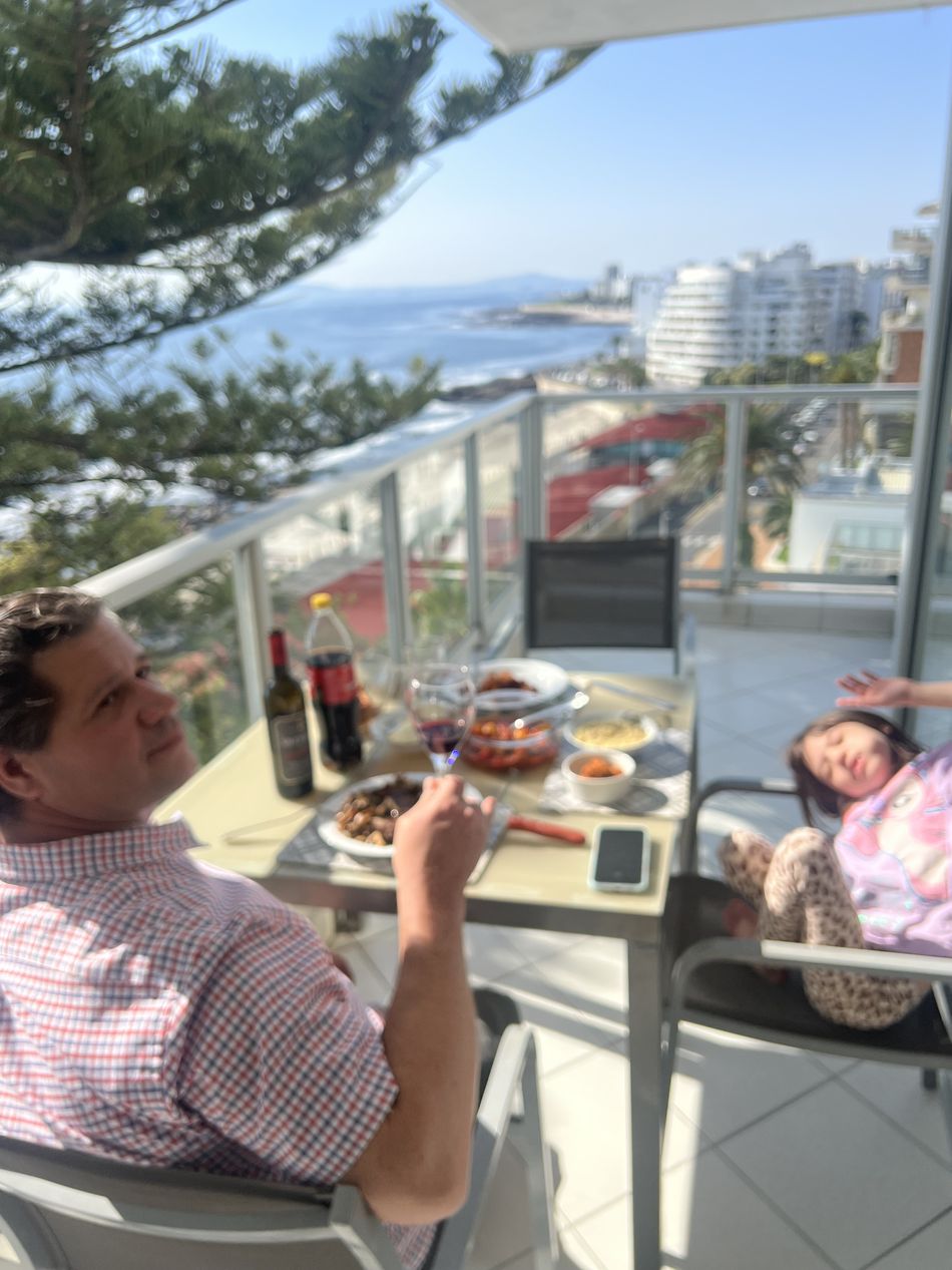 O Nicco almoçando na varanda do apartamento, com pratos e uma garrafa de vinho na mesa, o mar ao fundo e a Dinah esticada numa espreguiçadeira ao lado