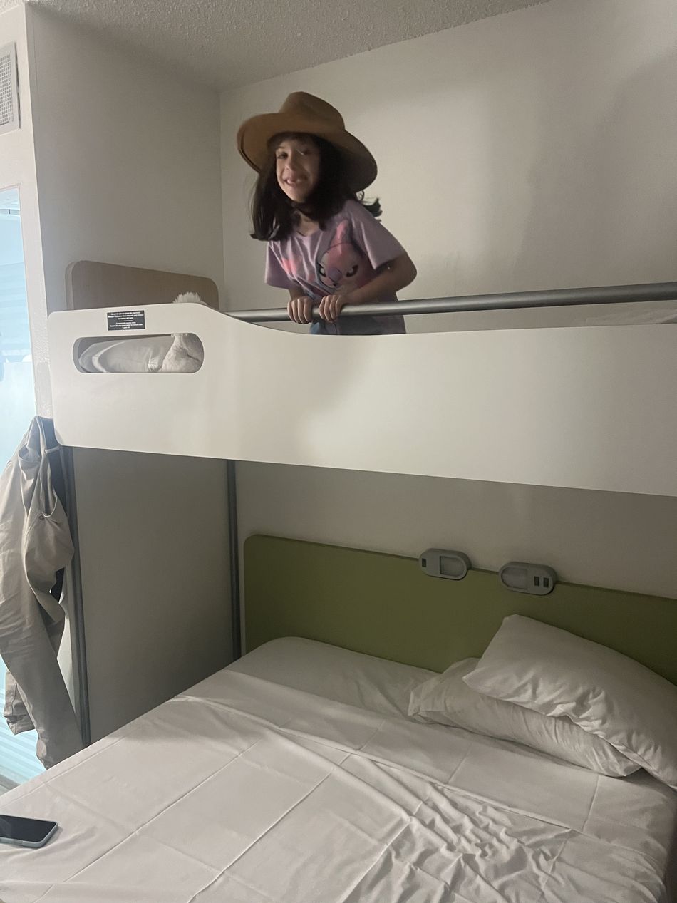 Dinah em pé no beliche superior do quarto do Ibis, de chapéu, sorrindo, com a cama de casal embaixo