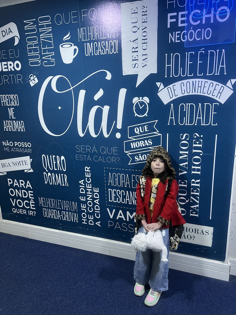 Dinah em pé diante do painel azul do hotel, coberto de frases em branco como Olá, quero um cafezinho e hoje é dia de conhecer a cidade
